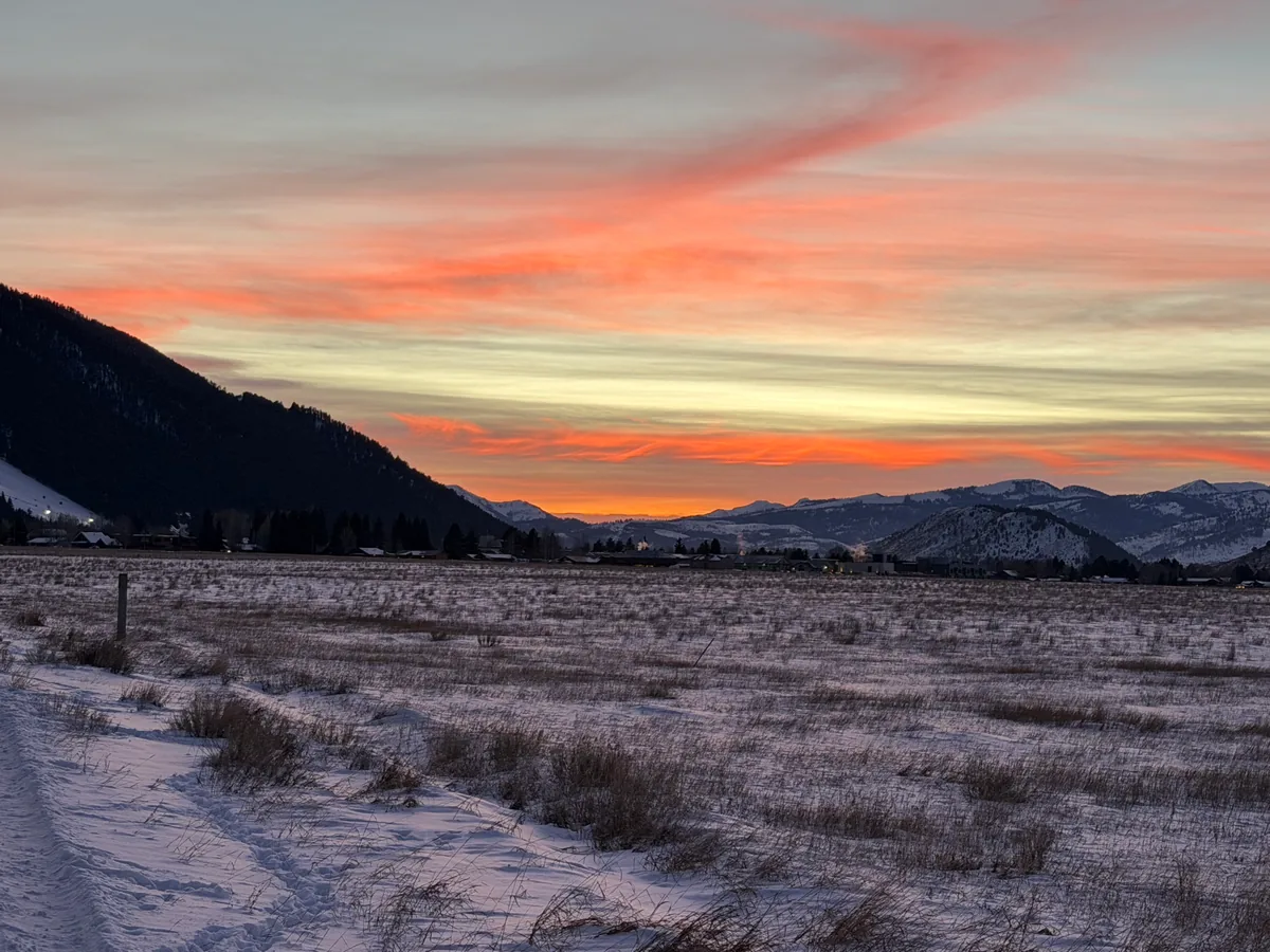 Orange and pink sunset clouds over snow-covered sage flats ringed by low dark mountains