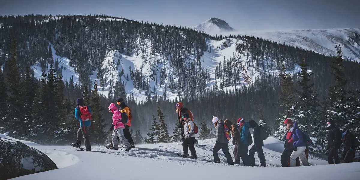 A long line of snowshoers in bright jackets crossing a snowy shelf below a wind-scoured peak