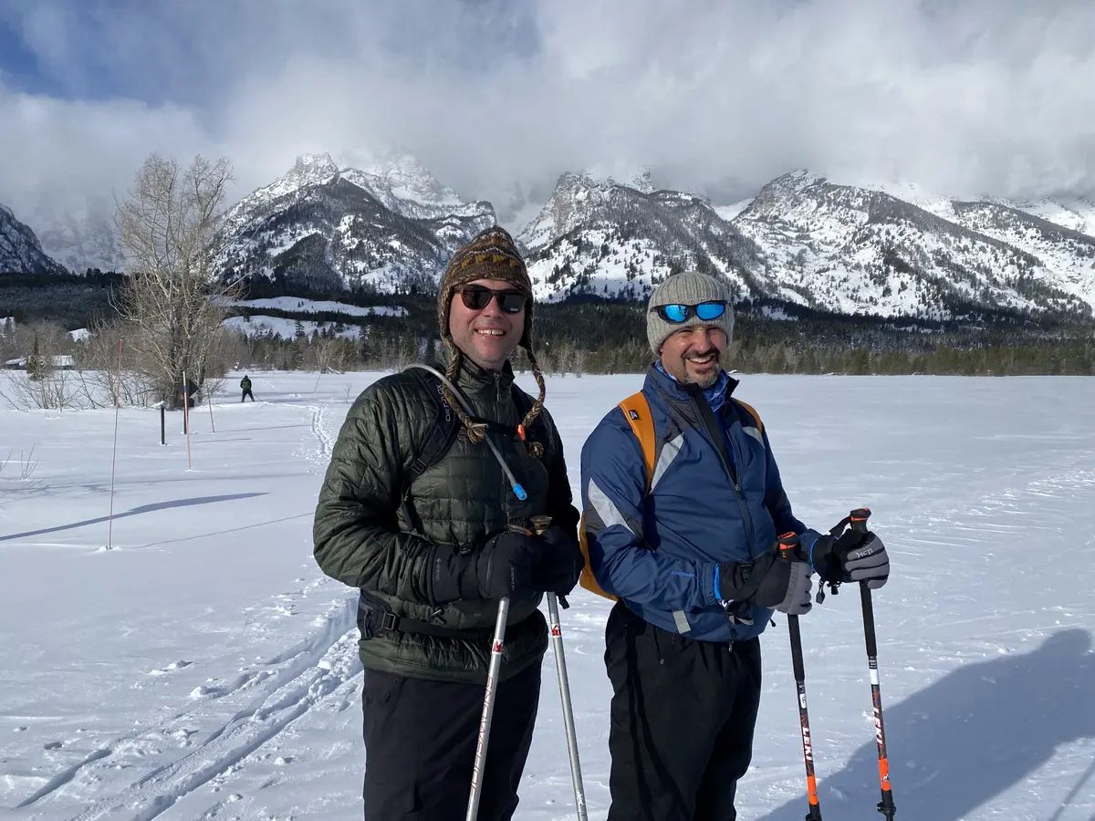 Two cross-country skiers smiling on an open snowfield beneath cloud-wrapped Teton peaks